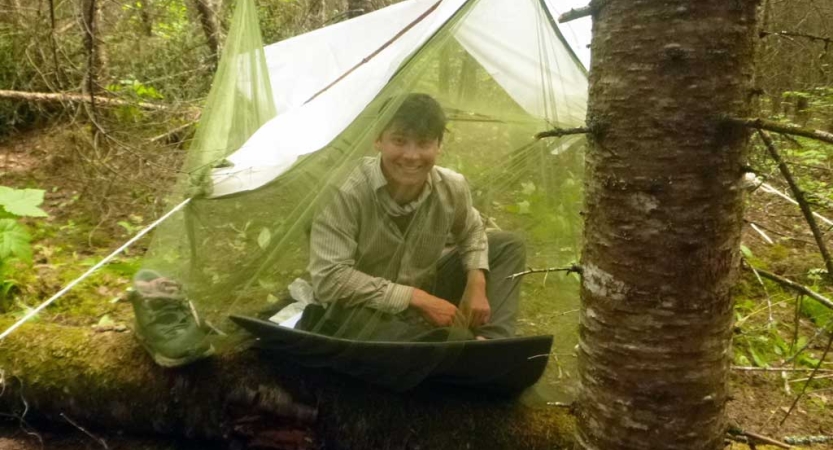 In a wooded area, a student sits and smiles inside a tarp shelter with a mosquito net draping it. 